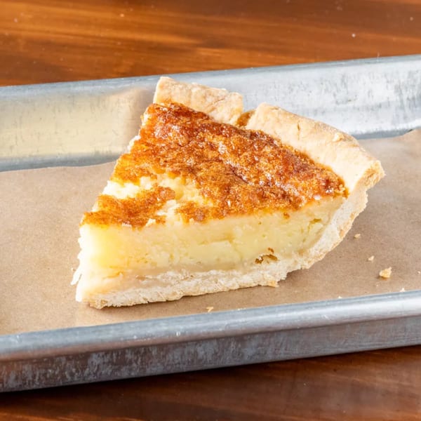 A slice of golden-brown custard pie with a flaky crust sits on parchment paper on a metal tray.