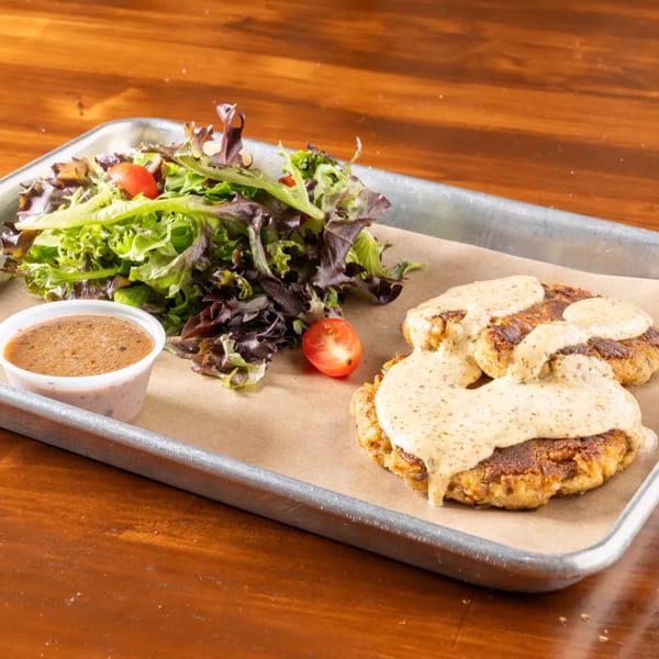 Two crab cakes with creamy sauce, a side salad, and dressing on a metal tray.